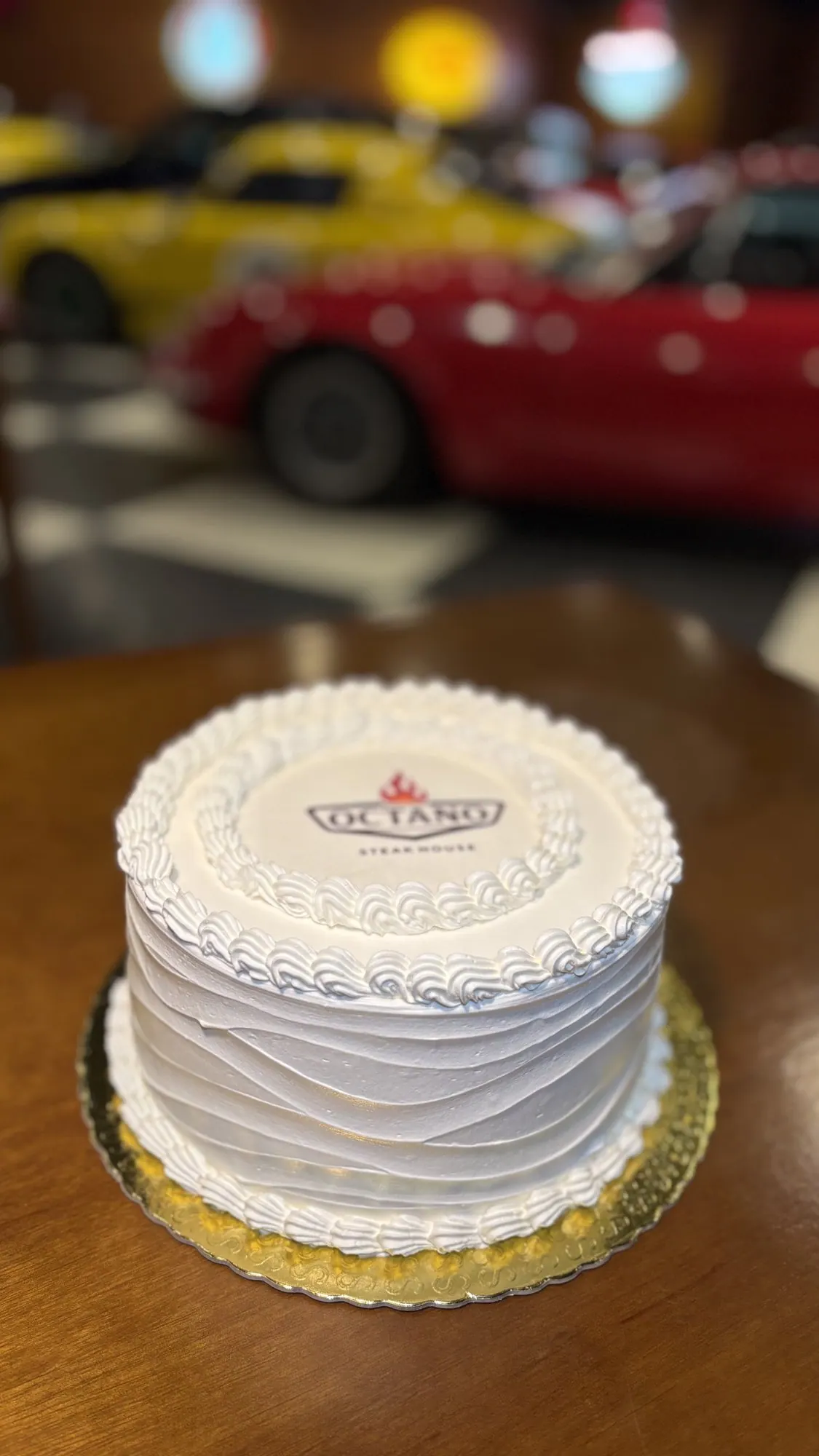 Bolo da casa com o logo do Octano, com os carros clássicos do salão ao fundo